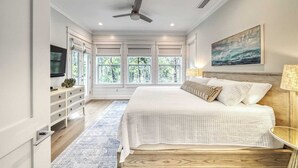 A bedroom with a large bed, a ceiling fan, a TV, and a window with a view of trees. - Amour La Mer by Avantstay Rooftop Patio, Pool Access, Walk to Beach (Panama City Beach)
