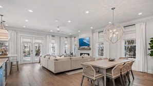 A spacious living room with a large sectional sofa, a dining area with a wooden table and wicker chairs, and a TV mounted on the wall. - Amour La Mer by Avantstay Rooftop Patio, Pool Access, Walk to Beach (Panama City Beach)