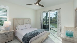 Un dormitorio con cama, mesita de noche con lámpara, ventilador de techo, ventana con cortinas y vistas a una piscina y palmeras.