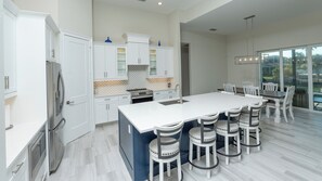Una cocina moderna con armarios blancos, una isla central con taburetes y vistas al exterior a través de amplias ventanas.
