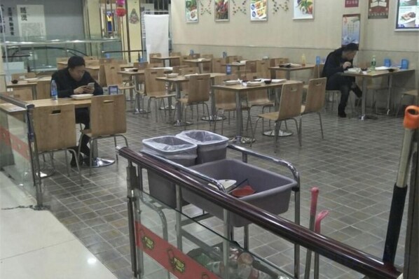 A cafeteria with multiple tables and chairs, a trash bin, and a person sitting at a table. - Shunhang Hotel (Harbin Taiping International Airport Branch) (Harbin)