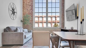 A modern living room with a large window, a sofa, a dining table, and a TV mounted on the wall. - Modern Escape | Pool | Easy Access to DT & More! (Greenville)