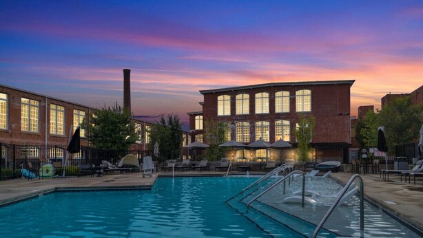 A swimming pool with lounge chairs and umbrellas, surrounded by brick buildings with large windows. - Modern Escape | Pool | Easy Access to DT & More! (Greenville)