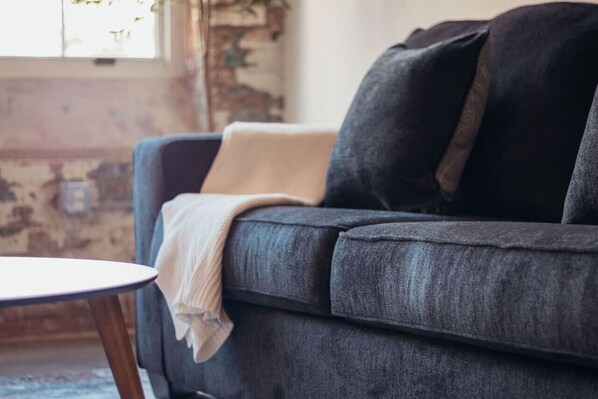 A dark gray sofa with a beige throw blanket and a pillow, positioned next to a small round table with wooden legs.