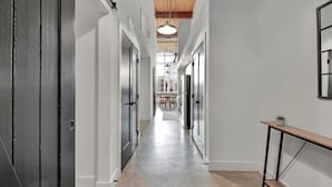 A hallway with white walls, a wooden ceiling, and a tiled floor. There is a table with chairs in the distance, and a small plant on a table to the right.
