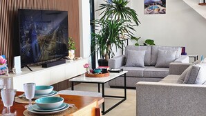 Ett modernt vardagsrum med en grå soffa, ett soffbord och en tv som är monterad på en vägg av trä. I bakgrunden syns växter, ett matbord med glas och en trappa.