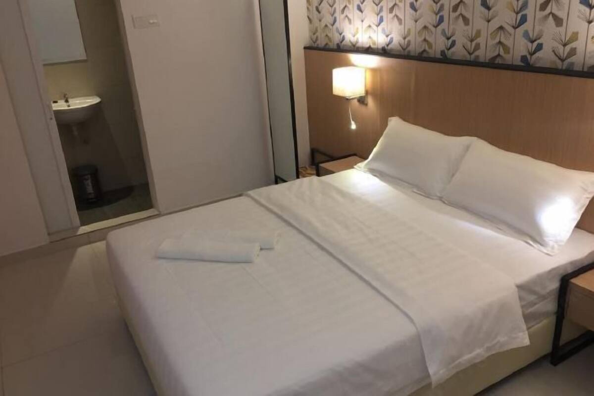 A hotel room with a bed, a nightstand, a wall-mounted lamp, and a bathroom visible through an open door.