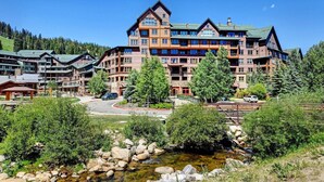 A multi-story lodge with a stone facade and multiple balconies, surrounded by lush greenery and a small stream.