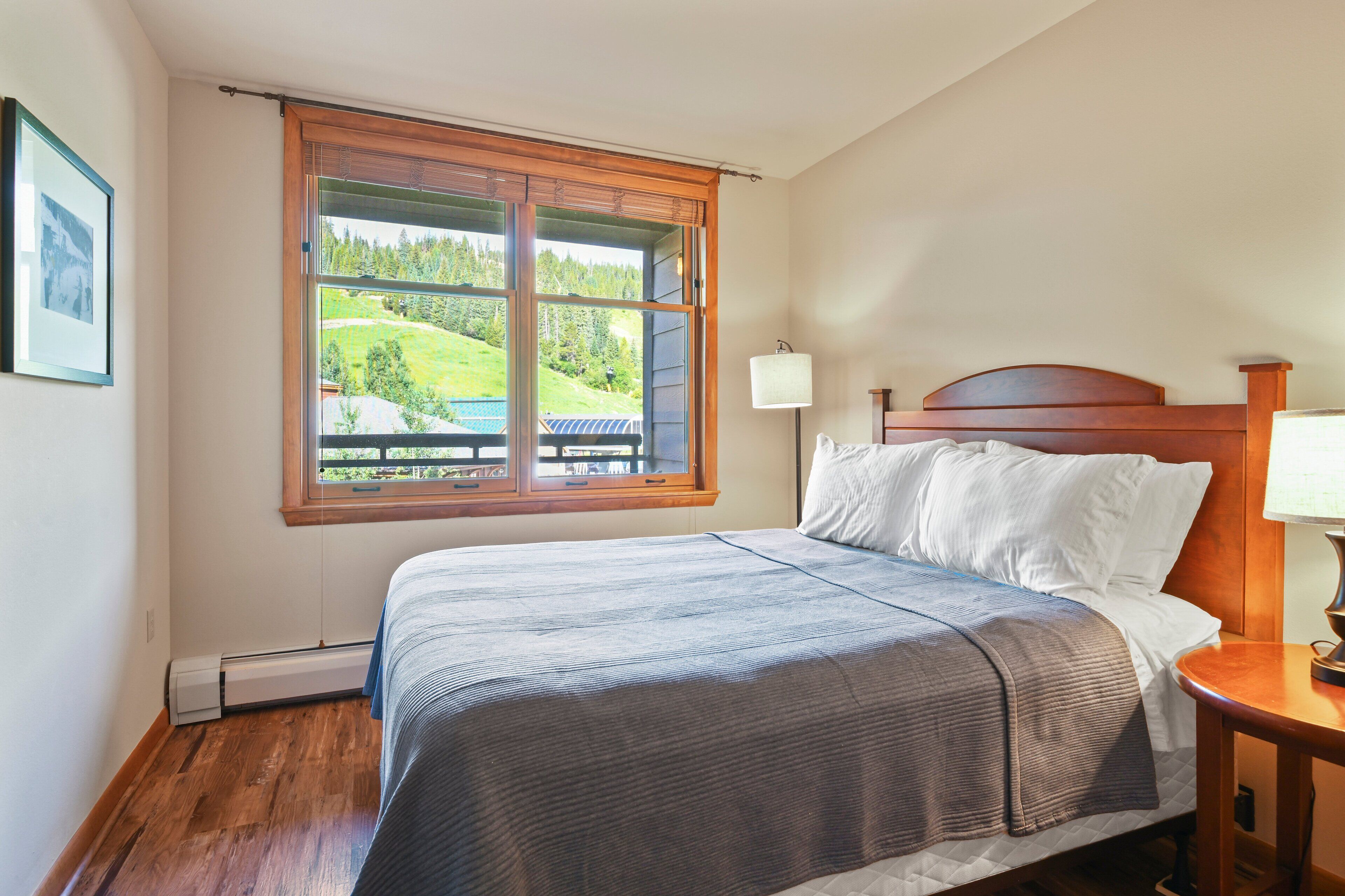 Un dormitorio con un ventanal grande, una cama con ropa de cama blanca, un cabecero de madera y una mesita de noche con una lámpara.