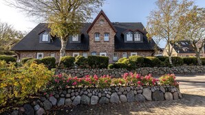 Een groot, twee verdiepingen tellend stenen huis met een steil dak, omringd door een stenen muur en bloeiende planten.