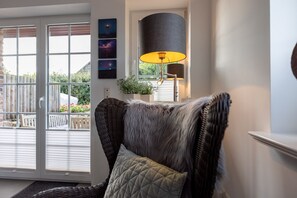 A cozy living room with a wicker chair, a lamp, and a view of a garden through a sliding glass door.