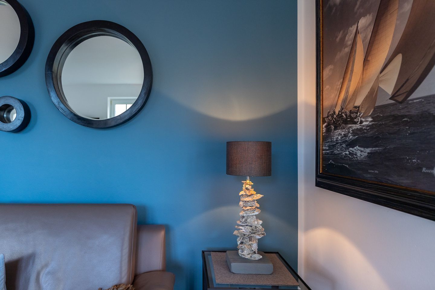 Quarto com parede azul, espelho redondo, pintura emoldurada, luminária com base diferenciada e poltrona de couro.