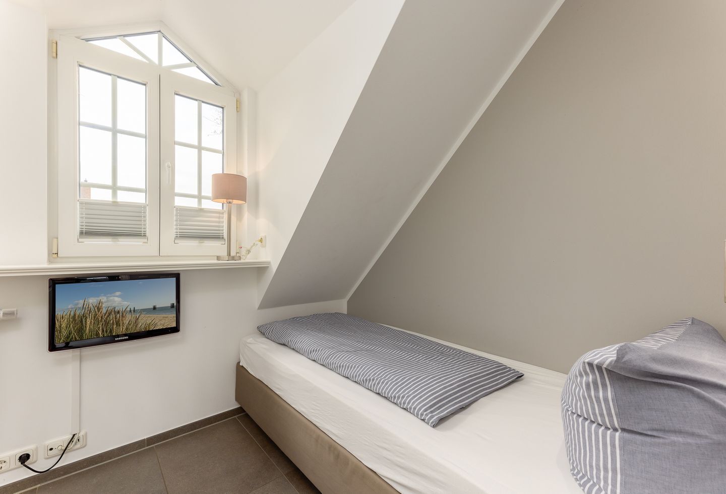 Une petite chambre avec un lit simple, un plafond en pente, une fenêtre avec des stores, une lampe de chevet et un téléviseur fixé au mur.