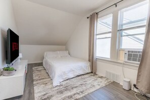 A bedroom with a bed, a TV, a window, and an air conditioning unit.