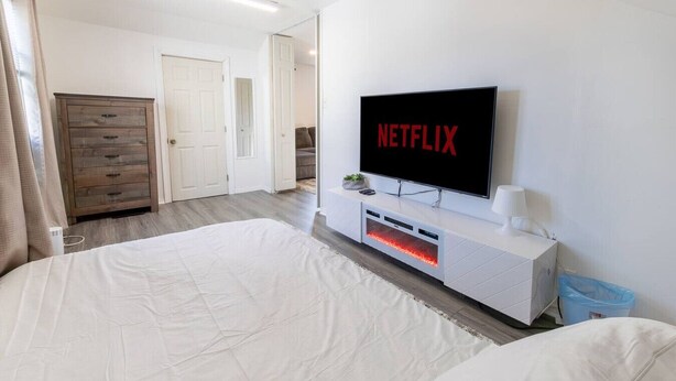 Una camera da letto con un letto grande, una TV a muro che mostra il logo di Netflix, un comodino bianco e un'armadio di legno.