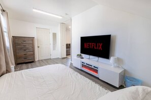 Ein Schlafzimmer mit einem großen Bett, einem an der Wand montierten Fernseher mit Netflix-Logo, einem weißen Konsolentisch und einem Holzschrank.