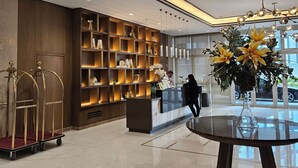 A modern hotel lobby with a reception desk, a glass-top table with a vase, a large window, and a wall of shelves with various items. - Luxury condo with gym,WiFi,NETFLIX at Park Mckinley West, Venice, SM Aura BGC (Taguig)