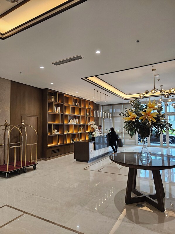 A modern hotel lobby with a reception desk, a glass-top table with a vase, a large window, and a wall of shelves with various items. - Luxury condo with gym,WiFi,NETFLIX at Park Mckinley West, Venice, SM Aura BGC (Taguig)