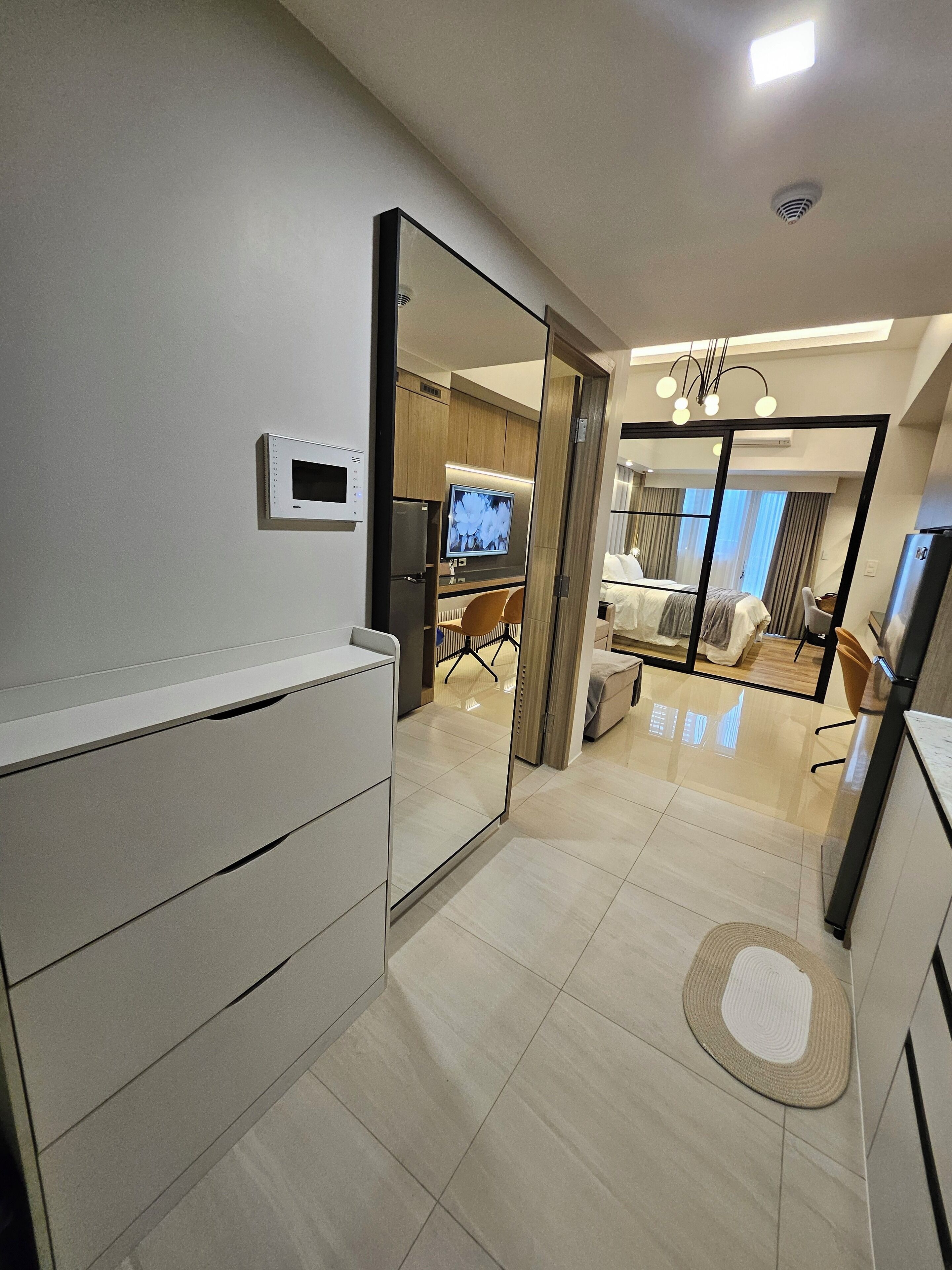 A modern hotel room with a large mirror, a flat-screen TV, a kitchenette, and a separate living area with a sofa and a dining table.