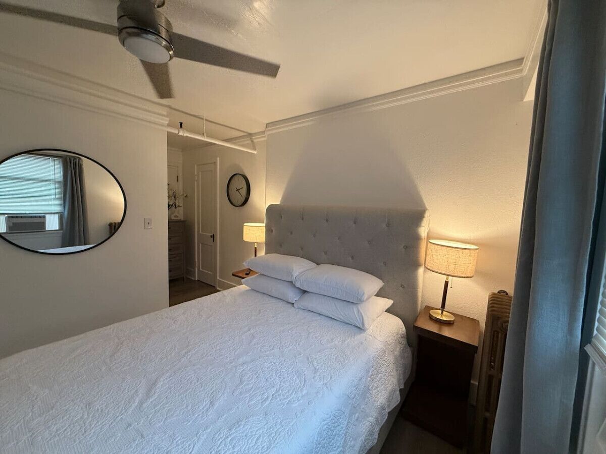 A bedroom with a bed, a round mirror, a clock, a ceiling fan, and a nightstand with a lamp.