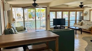 Ein großzügiger Wohnbereich mit Esstisch aus Holz, grünem Sofa und Fernseher. Große Fenster bieten Blick auf einen Pool und eine Außenanlage.