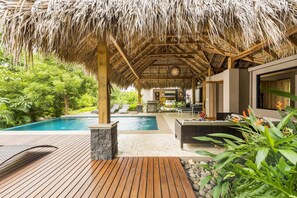 Un área de piscina con terraza de madera, una estructura de techo de paja y vistas a un exuberante jardín.