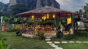 Un'area salotto esterna rustica con tetto di paglia, immersa in un giardino con sentieri in pietra che vi conducono.