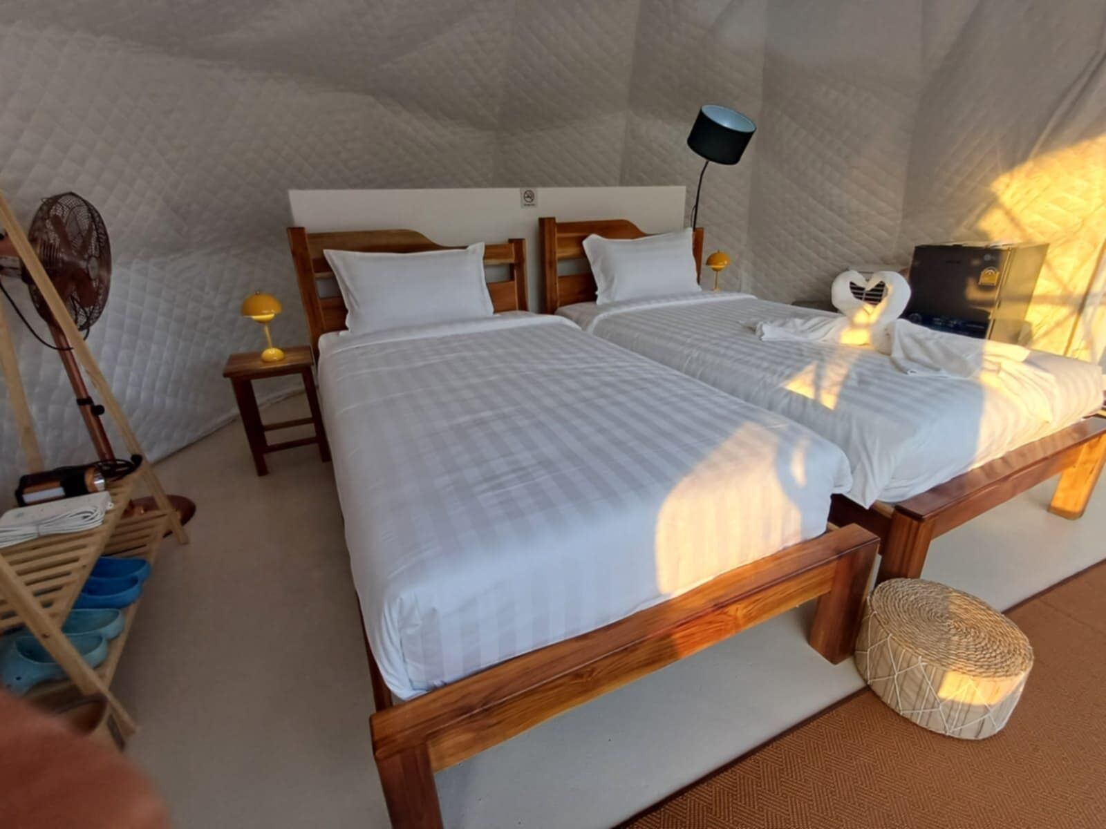 Una habitación acogedora con dos camas de madera, una mesita de noche con lámpara, un ventilador y un otoman tejido.