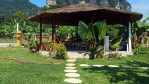 Un padiglione con tetto di paglia, raggiungibile tramite un viale in pietra, immerso nel verde lussureggiante e con montagne sullo sfondo.