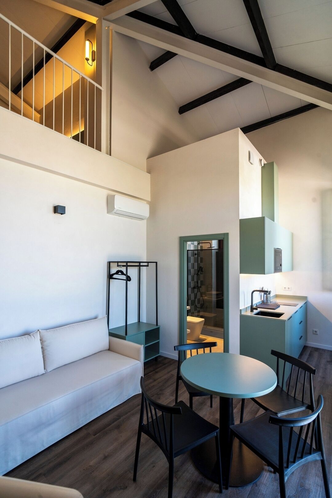 A modern interior with a sofa, dining table, and chairs. There is a staircase with a railing and a wall-mounted light fixture.