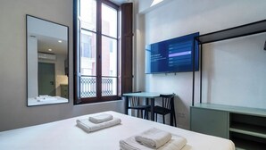A hotel room with a bed, a desk, a chair, a TV, and a window with a view of buildings.