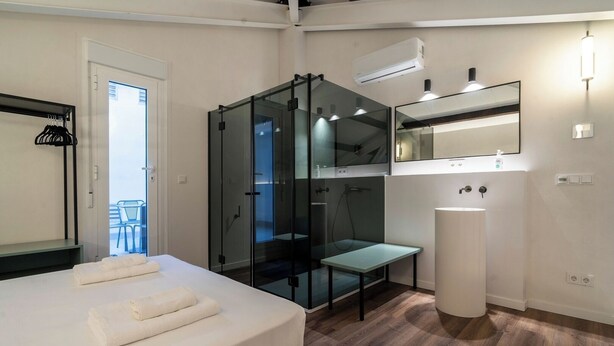 Habitación de hotel moderna con cama, zona de ducha con mampara de vidrio, una mesita, lavabo y balcón con sillas.