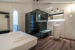 Moderni hotellihuone, jossa on sänky, lasiseinällä erotettu suihkutila, pieni pöytä, pesuallas ja parveke, jossa on tuoleja.