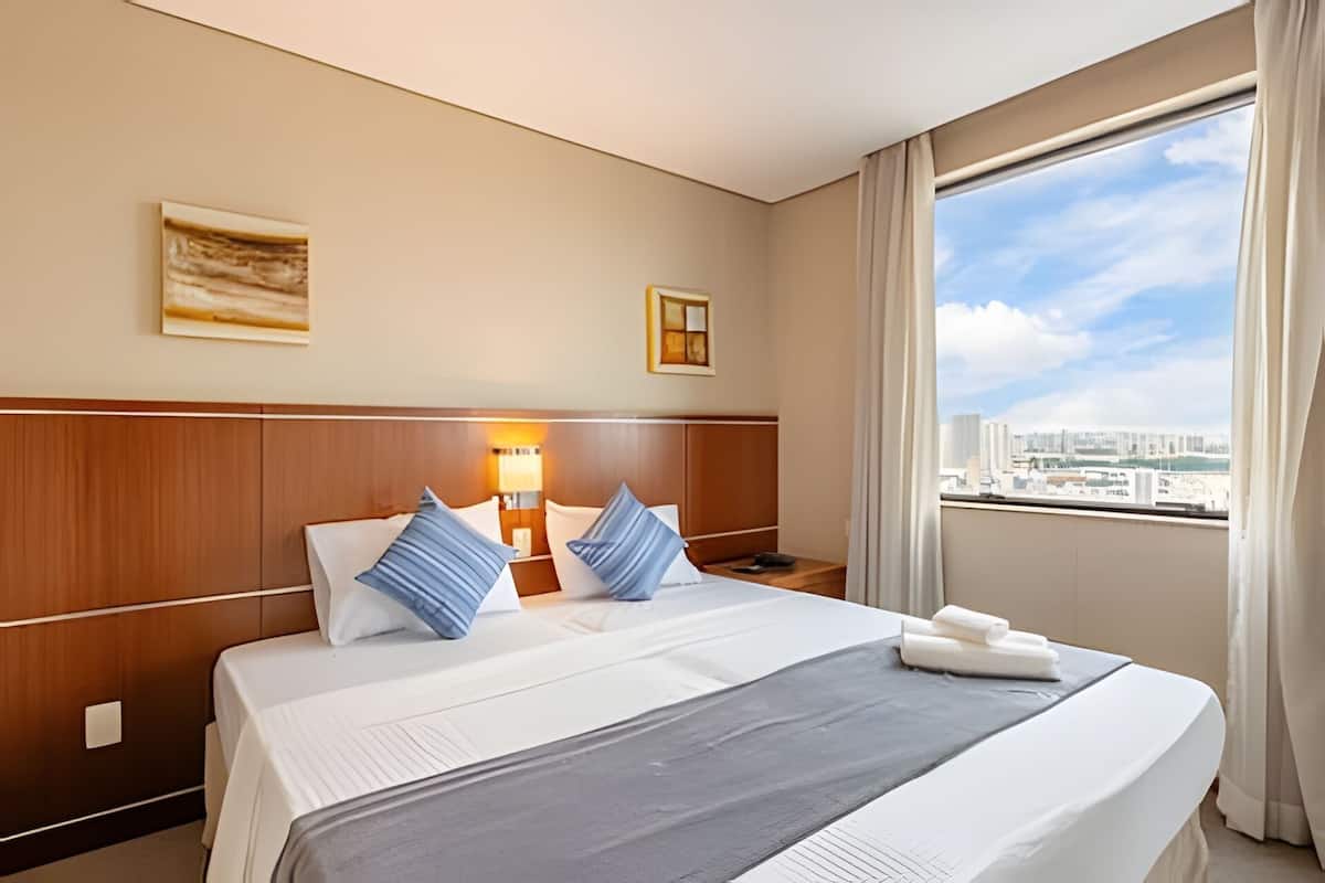 Ein Hotelzimmer mit einem großen Bett, einem Nachttisch, einer Lampe und Blick auf die Stadt.