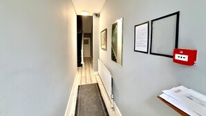 A narrow hallway with a wooden floor, a radiator, and a red emergency pull station on the wall.