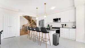 Une cuisine moderne avec des meubles blancs, un îlot central doté de tabourets de bar, des appareils en acier inoxydable et un escalier menant à un autre niveau.