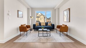 Un salon moderne avec un canapé bleu, deux fauteuils, une table basse et une grande fenêtre donnant sur un immeuble.