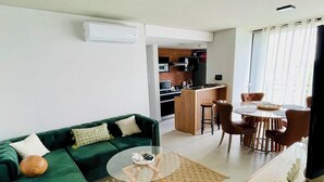 Een moderne woonkamer met een groene bank, een glazen salontafel en een eethoek met een houten tafel en stoelen.
