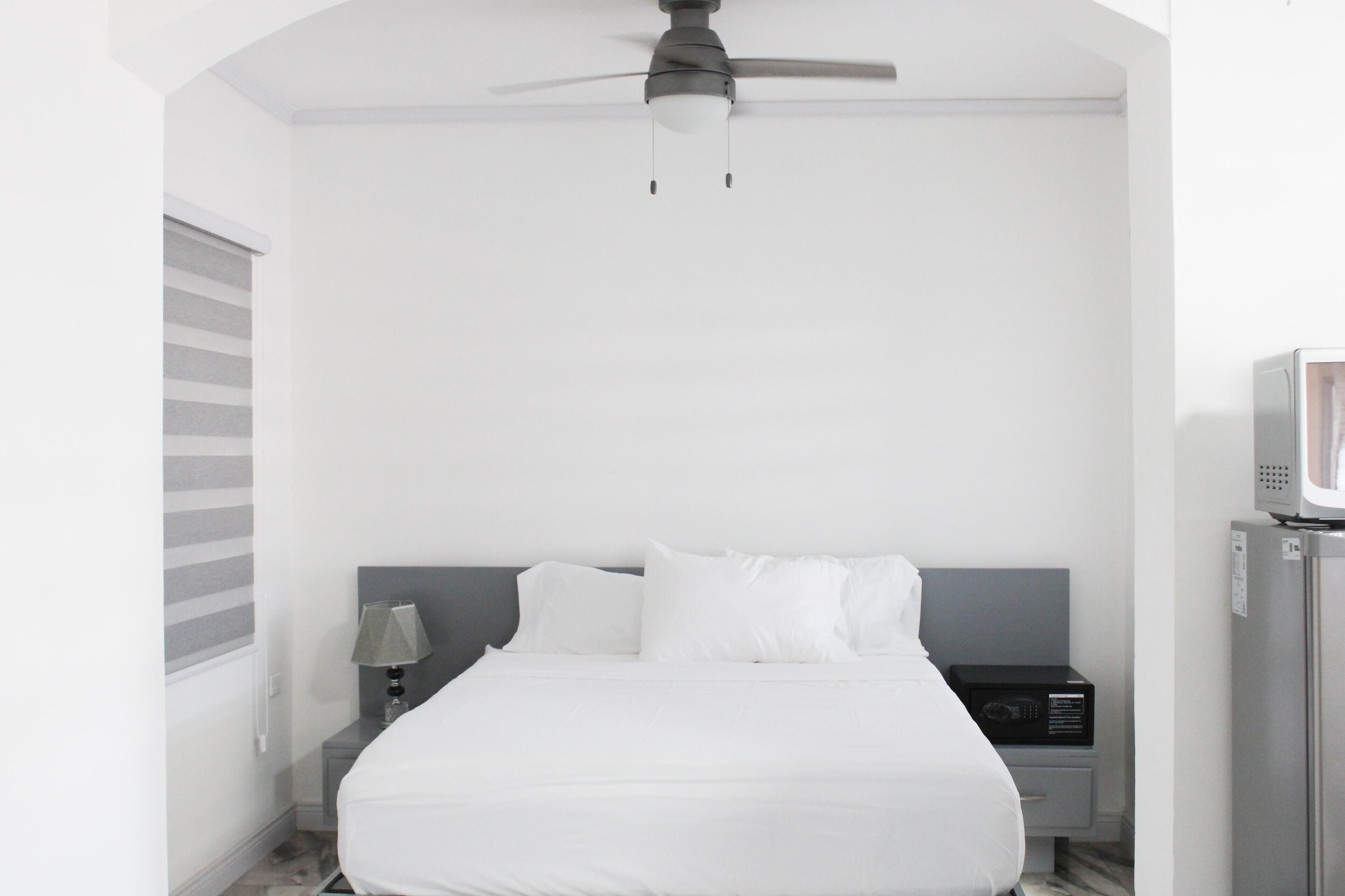 Una habitación de hotel con cama, mesita de noche, lámpara, ventilador de techo y microondas.