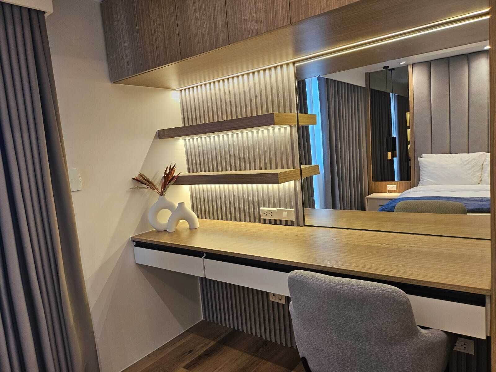 Ett modernt hotellrum med ett skrivbord i trä, en grå stol, en stor spegel och en säng med vita sängkläder.