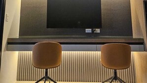 A modern kitchen with a flat-screen TV mounted on a wooden panel, two brown chairs with black legs, and a pendant light above the TV. - Luxury condo with gym,WiFi,NETFLIX at Park Mckinley West, Venice, SM Aura BGC (Taguig)