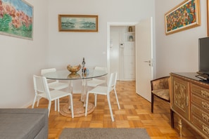 Uma sala de jantar com mesa de vidro e cadeiras brancas, uma televisão em um móvel de madeira, e uma porta que dá para um depósito.