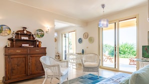 A spacious living area with a wooden cabinet, white wicker chairs, a blue patterned rug, and a sliding glass door leading to an outdoor patio.