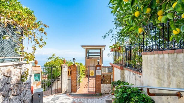 A stone staircase leading to a gated entrance with a metal gate, a small building with a glass door, and a view of the sea.