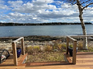 A wooden deck overlooking a calm lake with a rocky shoreline and a tree to the right. - Welcome to your relaxing, coastal getaway ! Oceanfront home nestled on the picturesque Orrs Island (Harpswell)
