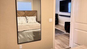 Ein Schlafzimmer mit einem großen Spiegel, einem Bett mit einer gesteppten Decke, einem Fernseher im angrenzenden Raum und einer Tür zu einem weiteren Bereich.