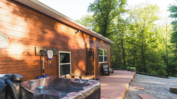 A wooden cabin with a hot tub, a covered patio, and a forested backdrop. - Bear's Den Retreat • Hot Tub • Soak Tub • Dog Friendly • Hocking Hills Cabin (Logan)