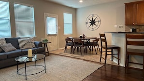 A living room with a sofa, coffee table, and dining table with chairs. There is a kitchen area with wooden cabinets and a stove.