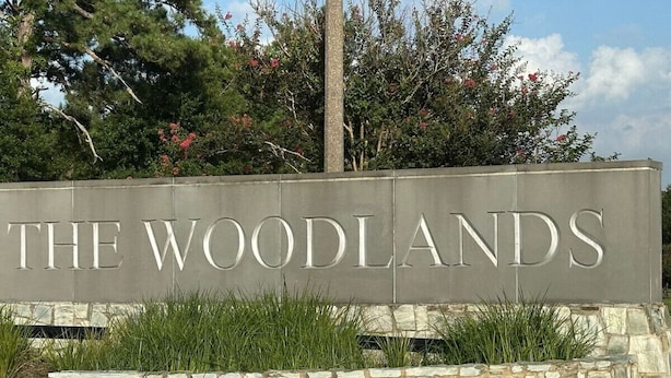 A sign for 'The Woodlands' with a stone wall and greenery in front.