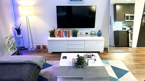 A modern living room with a flat-screen TV, a white entertainment unit with books and decorative items, a coffee table with a plant and a small sculpture, a sofa, and a blue LED strip light. - Downtown LA - Free Parking- Legends Loft! (Vernon)